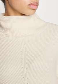 Donna che indossa un maglione a collo alto in maglia a coste color crema, mostrata dalla parte del collo fino alla parte superiore del torace, labbra visibili ma occhi fuori dal campo.