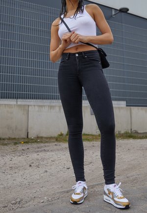 Osoba nosząca biały krótki top, czarne dopasowane jeansy, złoto-białe sneakersy, trzymająca czarną torebkę na ramię na zewnątrz obok szarej ściany.