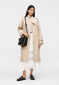 Γυναίκα που στέκεται φορώντας μια μπεζ παλτό τύπου trench πάνω από μια λευκή φούστα με δαντέλα, μαύρες μοκασίνια και κρατώντας μια μαύρη δερμάτινη τσάντα στον ώμο.