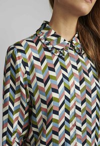 Chemise à manches longues avec un col à volants, arborant un motif en chevrons bleu marine, olive, rose et bleu clair sur un fond crème.