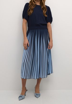 Femme vêtue d'un haut bleu marine à manches bouffantes, d'une jupe midi plissée à rayures bleues et noires, et de talons pointus bleus avec des détails en nœud.