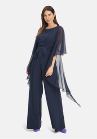 Marineblauer Jumpsuit mit fließendem Chiffon-Cape, tailliertem Bund, weitem Bein und lila Absatzschuhen. Glattes Material und eleganter Fall.