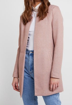 Vrouw draagt een lichtroze open mantel over een witte coltrui gecombineerd met hooggetailleerde blauwe jeans tegen een effen achtergrond.
