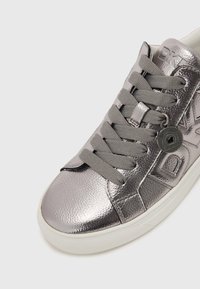 Tenis metalici argintii cu piele texturată, șireturi gri, detaliu logo modelat și talpă din cauciuc albă, vizibile dintr-o vedere unghiulară.