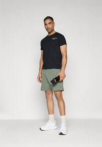 Zwarte T-shirt met korte mouwen, groene shorts, witte sneakers en een zwarte waterfles. Eenvoudig ontwerp, casual pasvorm en minimale details.