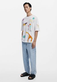 Witte graphic t-shirt met kleurrijke dierenillustraties en tekst. Gecombineerd met loszittende, lichtblauwe jeans met geborduurde zondetails. Zwarte sandalen.