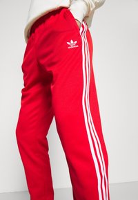 Červené sportovní kalhoty z hladké látky s bílými bočními pruhy a vyšívaným logem. Elastický pas s bočními kapsami.
