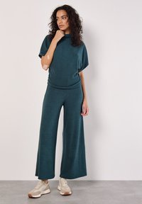 Teal tvådelad outfit med korta ärmar, avslappnad topp och vida byxor. Tyget verkar mjukt med en slät struktur, bärs med beiga sneakers.