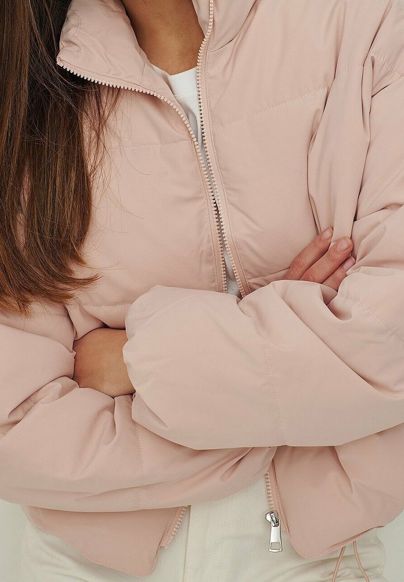 Personne portant une doudoune rose clair avec une fermeture éclair, les bras croisés, et de longs cheveux bruns visibles sur le côté gauche.