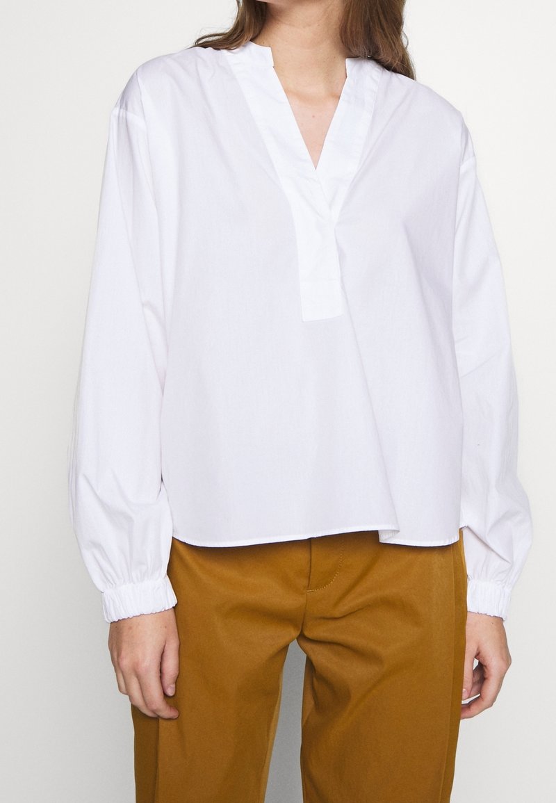 Vit långärmad skjorta med V-ringning och lös passform. Har samlade manschetter och en slät, krispig textur. Matchad med bruna, skräddarsydda byxor.