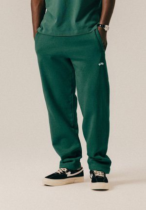 Osoba nosząca ciemnozielone spodnie dresowe z napisem "BLANC" na udzie, czarno-białe sneakersy, ręce w kieszeniach oraz zegarek na nadgarstku.