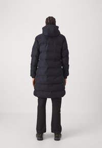 Lungo cappotto imbottito nero con cappuccio, caratterizzato da un design trapuntato, texture elegante e lunghezza fino a metà coscia. Indossato con pantaloni neri e stivali.