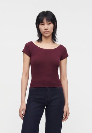 Jeune femme aux cheveux courts noirs, portant un haut bordeaux ajusté à manches courtes et un jean taille haute foncé, debout devant un fond uni.