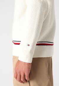Hvit strikket genser med tekstur, ribbet mansjetter og striper i rødt og marineblått nær nederste kant. Hender som holder fast i stoffet.