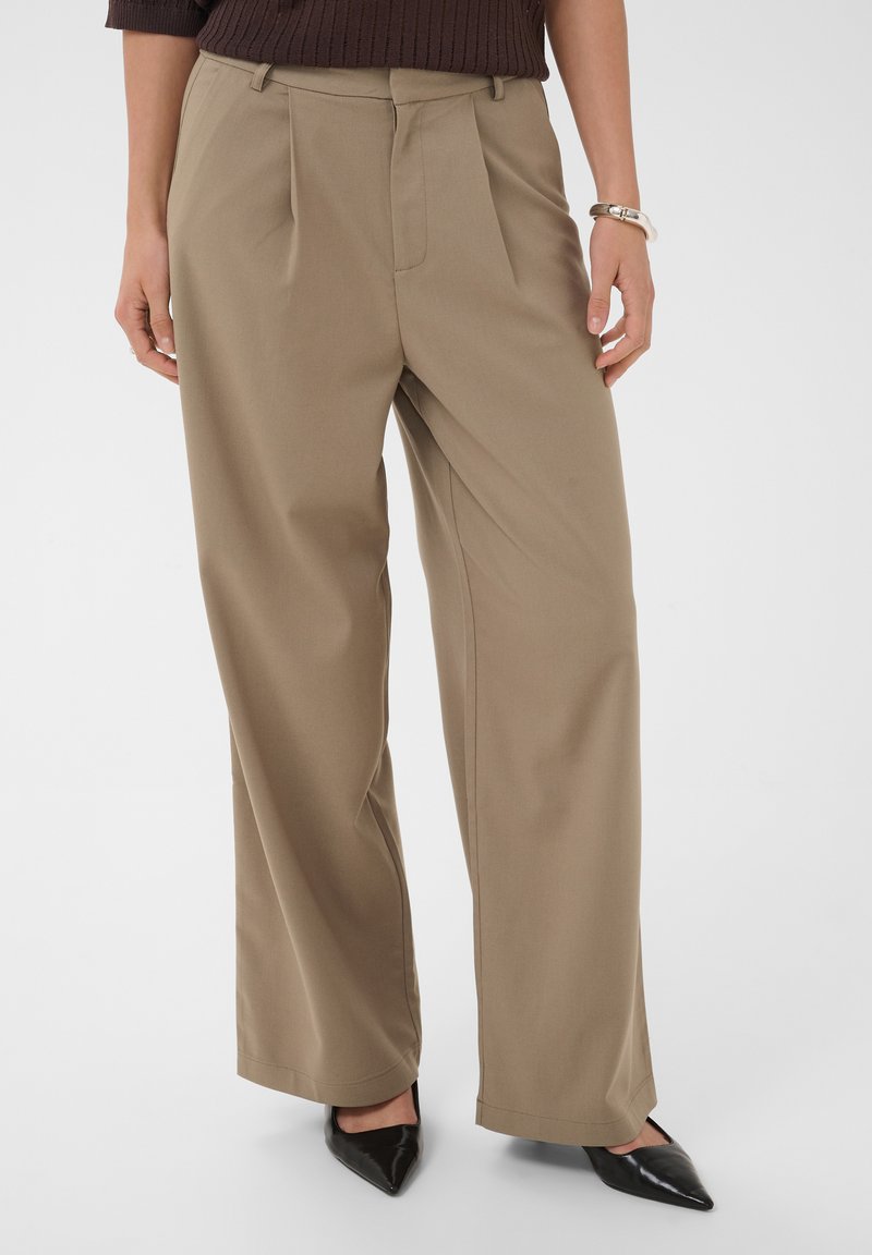Femme portant un pantalon beige à jambes larges, des chaussures noires à bout pointu et un pull côtelé marron, debout, mains détendues le long du corps.