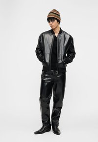 Άνδρας που φοράει μαύρο γυαλιστερό δερμάτινο μπουφάν και παντελόνι, μαύρες μπότες, ριγέ πλεκτό σκουφάκι και μαύρα γυαλιά ηλίου, με τα χέρια στις τσέπες.