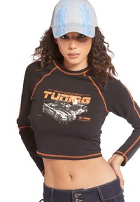 Maglietta nera a maniche lunghe cropped con dettagli arancioni, caratterizzata da una grafica di auto e la scritta "TUNING." Abbinata a un cappellino grigio chiaro con design di fiamme blu.