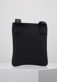 Černá kožená taška přes rameno s hladkou strukturou, která má dvě pevné rukojeti a minimalistický design. Ozdobena čtvercovými přezkami.