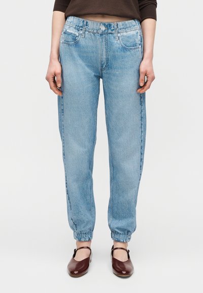 Άτομο που φοράει ανοιχτό μπλε τζιν παντελόνια τύπου jogger με ελαστικά μανσέτα και καφέ παπούτσια Mary Jane, πάνω σε απλό φόντο.