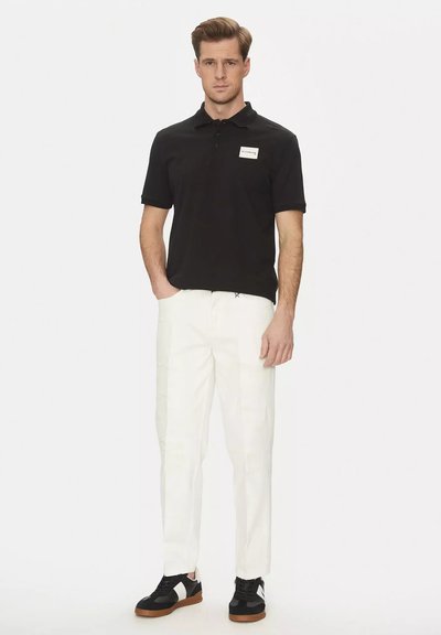 Maglietta polo nera con un patch sul petto, abbinata a pantaloni bianchi e sneakers nere con suole marroni, creando un outfit di contrasto.