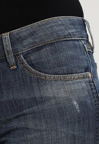 Modré džíny z denimu se přiléhavým střihem, klasickým pětikapsovým designem, diskrétním blednutím a cvoky na bocích.