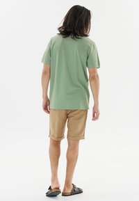 Grön kortärmad polo-shirt, beige rullade shorts och bruna sandaler. Skjortan har en mjuk textur och avslappnad passform. Enkelt design utan mönster.