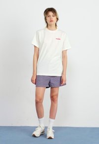T-shirt en coton blanc avec un petit logo rouge sur la poitrine, associé à un short rayé gris. Le mannequin porte des chaussettes blanches et des baskets claires.