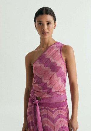 Mujer vestida con un top de un solo hombro y falda a juego con estampado de zigzag en rosa y púrpura, con un fajín atado en la cintura, de pie sobre un fondo liso.