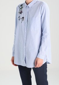 Camicia a maniche lunghe a righe blu chiaro con dettagli floreali ricamati, colletto classico, chiusura frontale con bottoni e taschino sul petto. Tessuto morbido.