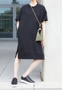 Vestido camisón negro de gran tamaño con mangas cortas y aberturas laterales. Accesorizado con un pequeño bolso de mano verde oliva y zapatillas negras.