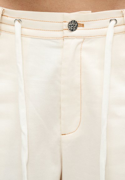 Κοντινή λήψη της μέσης σε μπεζ παντελόνι με μεταλλικό κουμπί, ορατό κείμενο μάρκας, λευκά κορδόνια και καφέ λεπτομέρειες ραφής.
