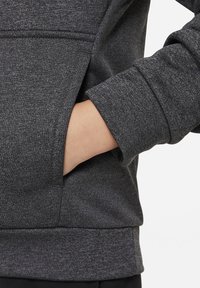 Felpa grigio scuro con tessuto strutturato, dotata di una tasca frontale e polsini a costine. Primo piano sulla manica e sulla mano nella tasca.