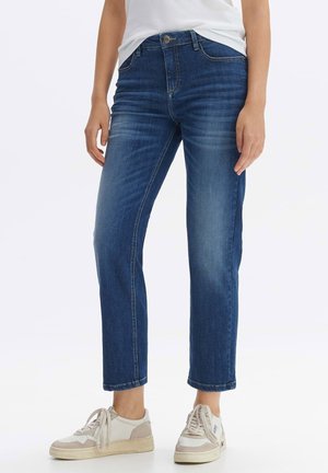 Femme portant un jean bleu taille mi-haute, coupe droite, avec des détails délavés, associé à des baskets blanches décontractées sur fond blanc.