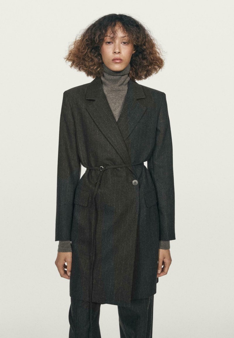 Mørkegrå, stribet blazer med slå-om design, knaplukning og matchende bælte. Stylet over en grå højhalset trøje, hvilket giver et lagdelt look.