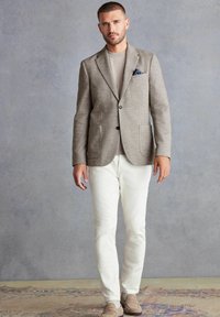 Hellbrauner, texturierter Blazer, beiges Hemd, weiße Hose, mit zwei vorderen Taschen und einem gefalteten blauen Taschentuch. Die Schuhe sind hellbraune Loafers.