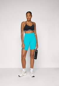 Sutien sport Nike negru cu logo, pantaloni scurți înalți turcoaz, adidași albi și o sticlă de apă neagră, stând pe un fond deschis.
