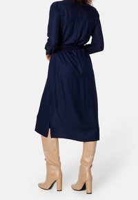 Abito lungo blu navy con maniche lunghe e vita cintrata, dotato di uno spacco sul retro. Indossato con stivali beige al ginocchio, realizzati in pelle testurizzata.