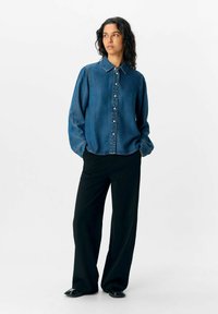 Blå denimskjorta med knappar, krage, långa ärmar och en avslappnad passform, i kombination med vida svarta byxor och blanka svarta skor.