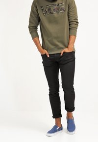 Sudadera con capucha verde oliva con un logo de camuflaje, jeans negros de corte ajustado y zapatos azules tipo slip-on. Conjunto casual, diseño minimalista.
