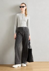 Haut gris à manches longues avec patte de boutonnage, associé à un jean noir à jambes larges. Des baskets blanches épaisses et un sac fourre-tout noir complètent la tenue.