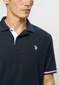 Tummansininen poolopaita, jossa on valkoinen nappilista, lyhyet hihat, joissa on punaisia ja valkoisia raidoituksia, sekä brodeerattu logo rinnassa.