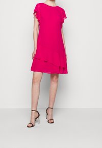 Fuchsia pink dress with short ruffled sleeves and layered design. Features a rounded neckline and falls above the knees. Worn with black strappy heels.