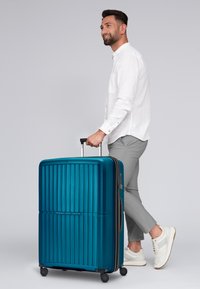 Blauer Hartschalenkoffer mit vertikalen Rillen, ausziehbarem Griff, vier Rollen und einem schlanken Design. Glatte Textur mit glänzender Oberfläche.