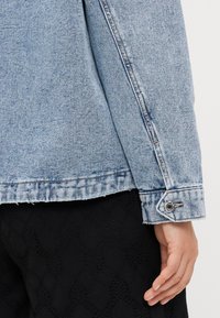 Lichtblauwe spijkerjasje met onafgewerkte zoom, knoopmanchetten en doorgestikte details; gedragen over een zwart textuur kledingstuk.