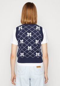 Navyblauer Strickwesten mit einem weißen Diamantenmuster und Schleifen. Die Weste hat einen gerippten Saum und wird über einem weißen T-Shirt getragen.