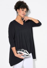 Schwarzes Langarmoberteil mit V-Ausschnitt und Falten-Detail. Das Model hält eine metallische Lippen-förmige Clutch und trägt dazu weiße Hosen.