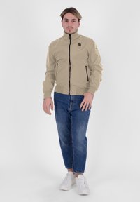 Giacca beige con zip e colletto alto, due tasche laterali e dettagli neri sulla zip, indossata sopra jeans blu e sneakers bianche.
