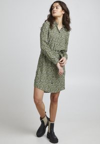 Abito floreale verde con maniche lunghe, chiusura a bottoni e cintura da annodare. Abbinato a stivali neri alla caviglia con dettagli beige. Tessuto liscio.