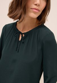Blusa verde scuro in tessuto leggero con scollatura arricciata, dettaglio a occhio di bue e inserti con laccetti regolabili. Maniche lunghe con una morbida texture.
