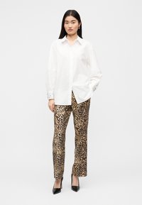 Λευκό πουκάμισο με κουμπιά και μακριά μανίκια με λεπτομερείς μανσέτες, συνδυασμένο με παντελόνι με σχέδιο λεοπάρ. Η μοντέλο φοράει μαύρες μυτερές γόβες.